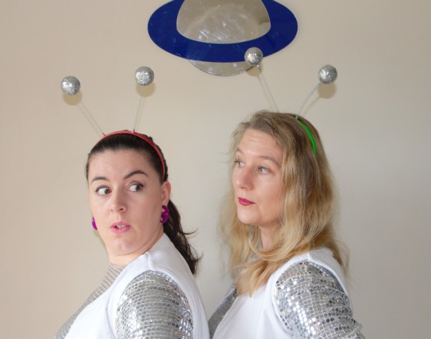Head and shoulders view of two women in profile wearing novelty headbands. One looks surprised. They both wear silver costumes.