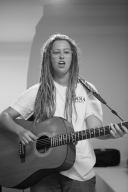 Black and white. A woman with long blond dreadloacks, wearing a white t-shirt plays the guitar and sings.