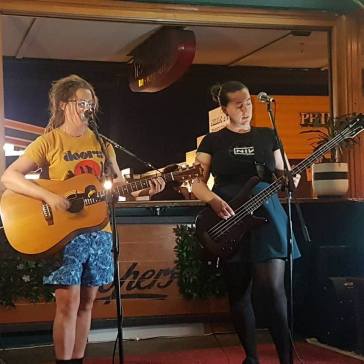 Two women perform in a bar. The woman on the left has blond dreadlocks and holds a guitar, the woman on the right has brown hair and holds a bass guitar.